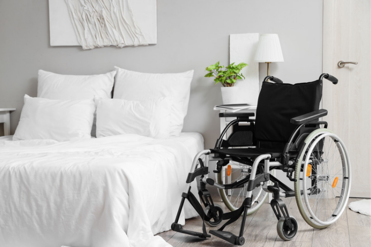 Wheelchair positioned beside a neatly made white bed in a modern, accessible bedroom. [Image by pixelshot on Canva.]