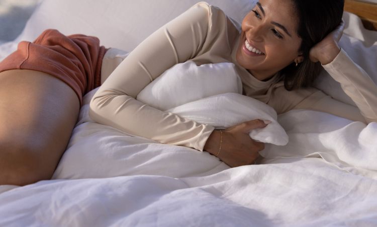 A woman is lying on a bed, smiling and hugging a white pillow.