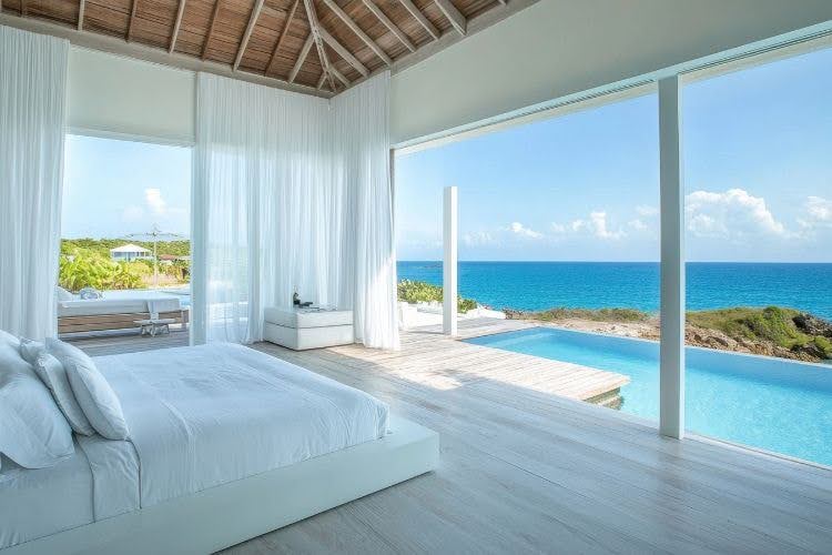 Minimalist white bedroom overlooking a pool and ocean with open curtains and wooden flooring. [Image by anytka on Canva.]