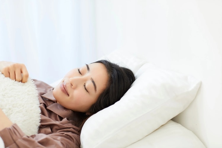 Woman resting peacefully on white pillows and bedding in a bright, minimalist room. [Image by Aflo Images on Canva.]