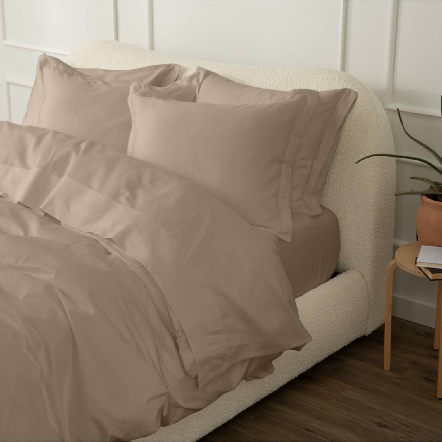 Beige bedding on a white upholstered bed with a plant on a side table.
