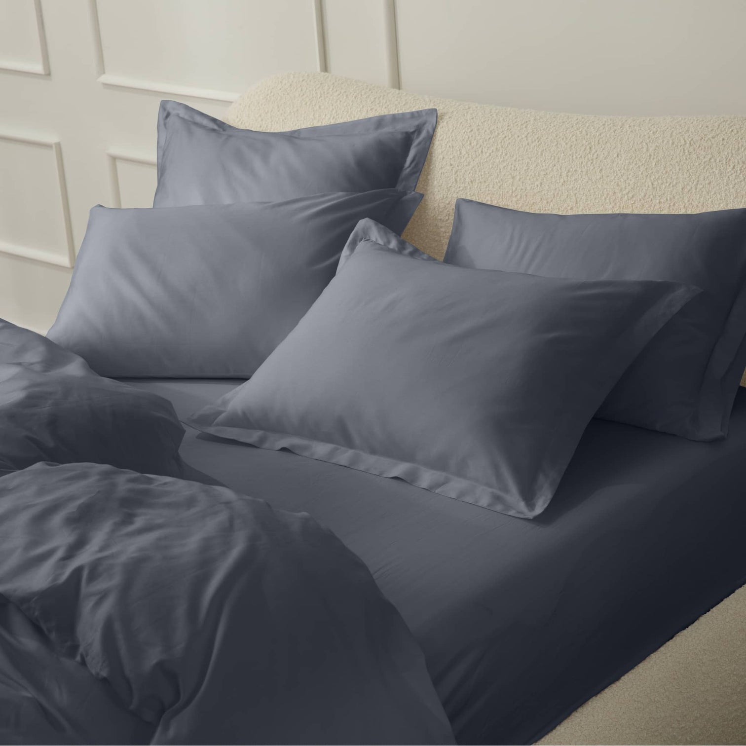A bed with gray sheets and multiple pillows against a light-colored wall.